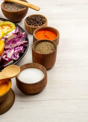 bottom-view-cut-vegetables-fruits-pumpkin-bell-peppers-persimmon-red-cabbage-black-plate-spice-small-bowls-wooden-spoon-wooden-table-copy-space